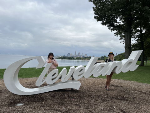 Two Queer Peer Mentors mentees stand behind the Cleveland script sculpture with the Cleveland skyline in the background