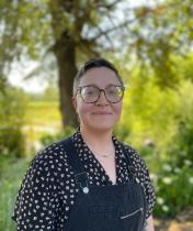 Mailey is a white person with short cropped brown hair and glasses. they are wearing black overalls and a black button up with small pink flowers printed on it. They are smiling in front of a lush outdoor background