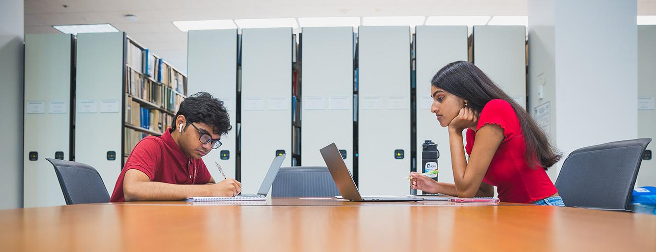 Two students work on computers at a table in front of the stacks at Kelvin Smith Library.