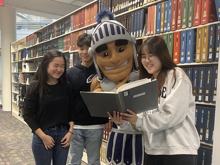Students and Spartie look at a book in front of a bookshelf at Kelvin Smith Library.