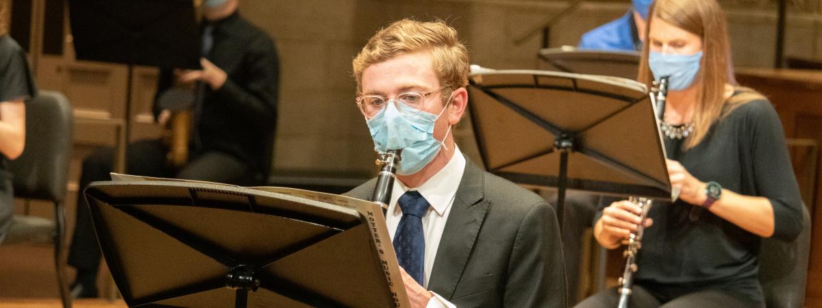 a man sits playing the clarinet with a mask on while a woman sits behind, also wearing a mask and playing the clarinet