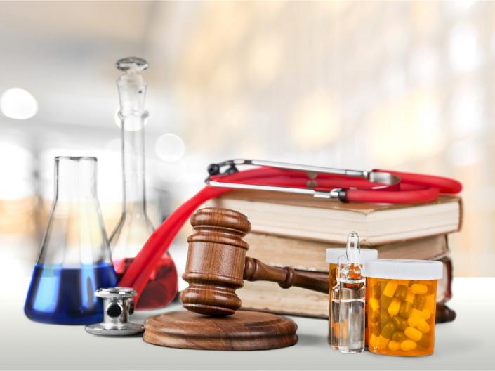 Various objects are piled together including several flasks filled with colored liquid, brown books, orange pill bottles, a stethoscope, and a gavel.