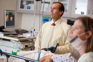 Image of 2 medical professionals with a patient lying between them, with the person in the center, dressed in a yellow surgical gown, is hoding a black tube-like instrument and they are both looking up at a screen, with a monitor and equipment in the background