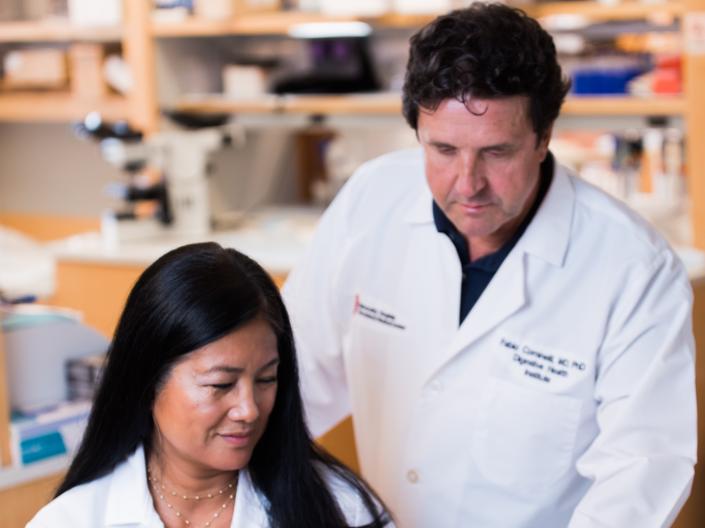 Image of 2 researchers in white lab coats looking at a white form in a medical setting with white shelves and some medical supplies in the background