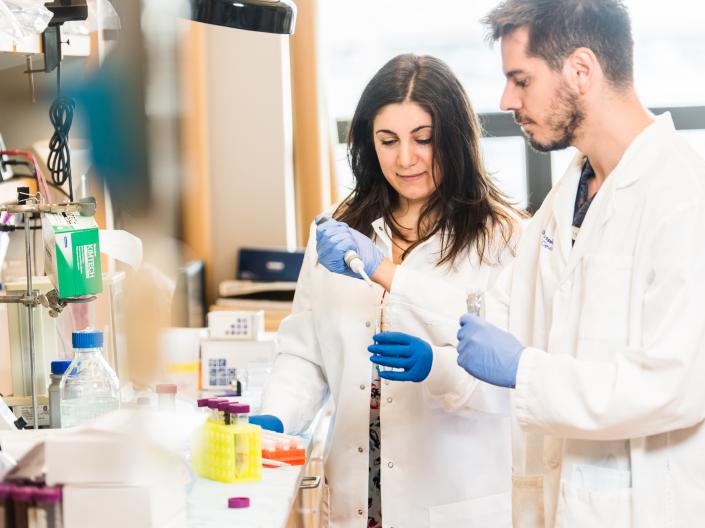 Image of 2 researchers in white lab coats looking at a medical tube in a medical setting with white shelves and some medical supplies in the background