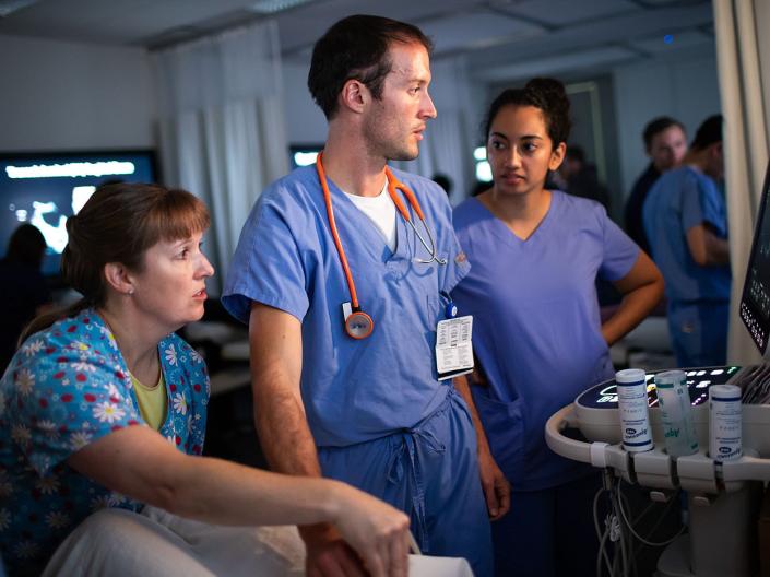 Three medical students in scrubs chat around medical equipment 