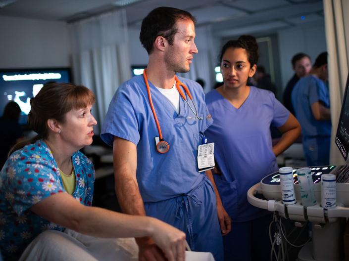 Three medical students in scrubs chat around medical equipment