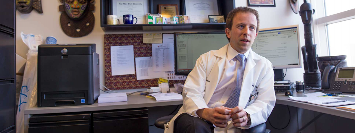 Image of doctor in his office.  He has short, brown hair, wearing a white lab coat, white dress shirt, and blue and white cross designed tie.  He is surrounded by a black file cabinet, black printer, two monitors, an african sculpture, and a phone.  On the wall behind him are two african masks, two degrees, and one award