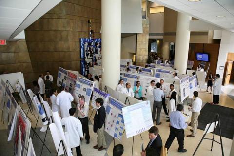 Image of large lobby with white pillars with about forty display posters on dispay tripods spread out in rows with about thrity people, half in white lab coats conversing and viewing the displays.  there is a white, blue and red mosiac design on a far wall and a display screen in blue on the another