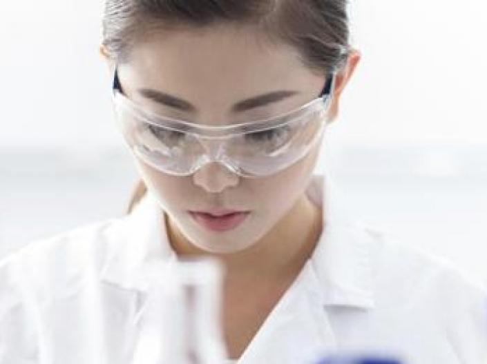 Image of researcher in white labe coat and safety goggles, looking down in analysis. There are the tops of three pieces of lab equipment, one white and two with blue tops, in the foreground