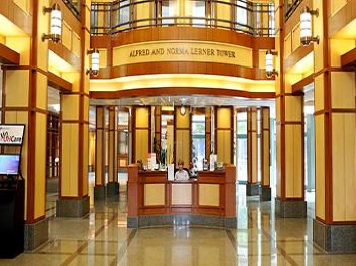 Image of lobby of Alfred and Norma Lerner Tower. The area is lit up with bright yellow lights, with small plants and kiosks on both sides. There is old fashioned lighting and a second floor with a railing above. The receptionist is seated in a wood panelled area in the center