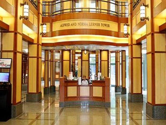 Image of lobby of Alfred and Norma Lerner Tower.  The area is lit up with bright yellow lights, with small plants and kiosks on both sides.  There is old fashioned lighting and a second floor with a railing above.  The receptionist is seated in a wood panelled area in the center