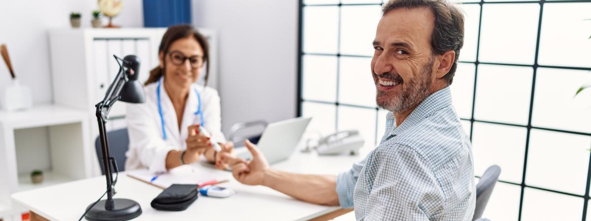 A middle-aged masculine patient smiles toward the camera as a femme and LatinX doctor measures measures his blood glucose