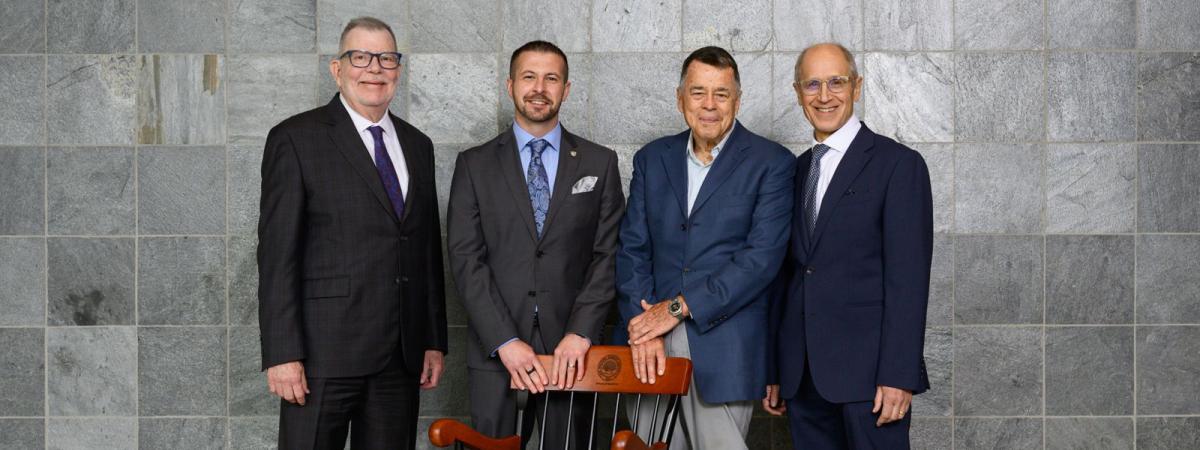 Four men in business attire stand side by side in front of a gray stone wall, posing for a formal photo. One man rests his hands on the back of a wooden chair positioned in front of them.