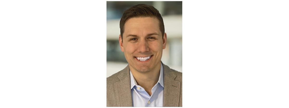 Professional headshot of Benjamin Clayton smiling toward the camera while wearing a light blue collared shirt and tan blazer. The portrait is set against a softly blurred indoor background with neutral tones.