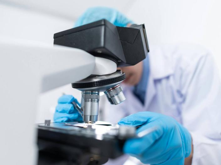  man in a lab coat examines a specimen through a microscope.