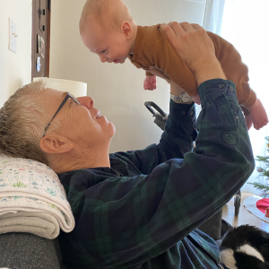 Man seated in a chair holding a baby above his head. Man and baby are smiling at each other.