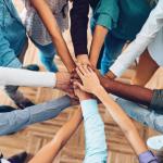 A picture of people standing in a circle extending their arms into the center of the circle so that their hands meet. Only the arms are visible.