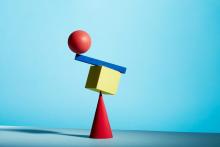 A cone shaped wooden block is supporting a square shaped block which is supporting a rectangular block which is supporting a round block. 