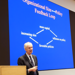 Photo of Larry Casalino standing on stage next to a podium and in front of a large screen projecting a slide titled Organizational Size [and] Policy Feedback Loop 