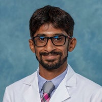 Man with dark hair, glasses, beard and mustache in white coat and tie