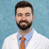 Man with dark hair, beard and mustache in white coat and tie