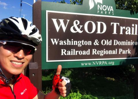 Male masters student wearing bike helmet in front of sign reading W&OD Trail