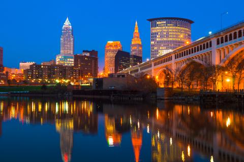 Downtown Cleveland skyline at dusk with a bridge on the left and the Cuyahoga River with skyline reflections below.