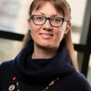 Portrait of Daria Fedyukina - woman with glasses wearing a dark sweater and a necklace standing in front of  a window. 