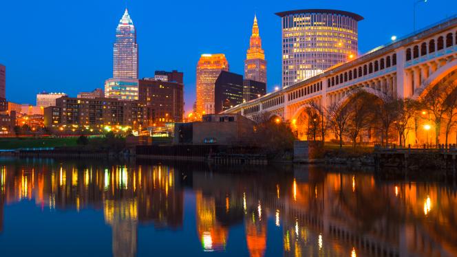 Downtown Cleveland skyline at dusk with a bridge on the left and the Cuyahoga River with skyline reflections below.