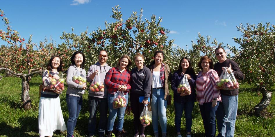 A group of 10 people stand in an apple orchard holding fresh-picked apples.