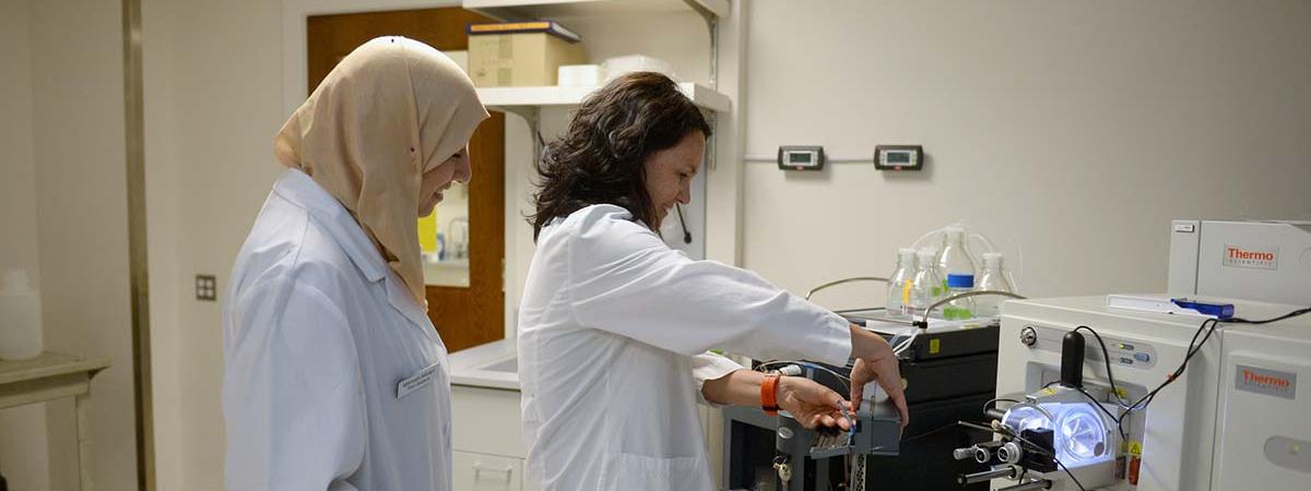 A woman wearing a lab coat demonstrates how to use a scientific instrument to a trainee.