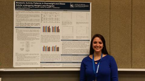 Image of Stephanie Logosh, in blue and wearning a blue lanyard around her neck, in front of display poster with text, bar and line graphs, all illegible except the title Metabolic Activity Pattersn in Overweight and Obes Adults undergoing Weight Loss Prograrm, and the logo in upper right of white square, with white lines shooting out of a clue circle, and the words school of medicine case western reserve university