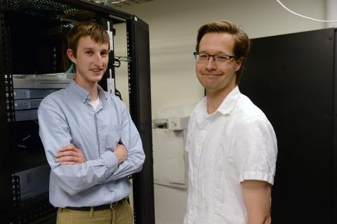 Two men smile at the camera while standing in front of a rack of computer equipment.