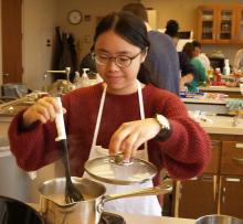 A student wearing a red sweater and glasses stirs a pot of rice.