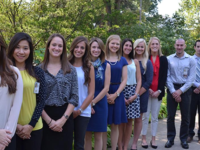 Image of twelve students in dress attire lines up in a row decending away from the camera smiling. they are on a concrete path in a green wooded area with a large tree behind the first person on the left. The sun is shining.