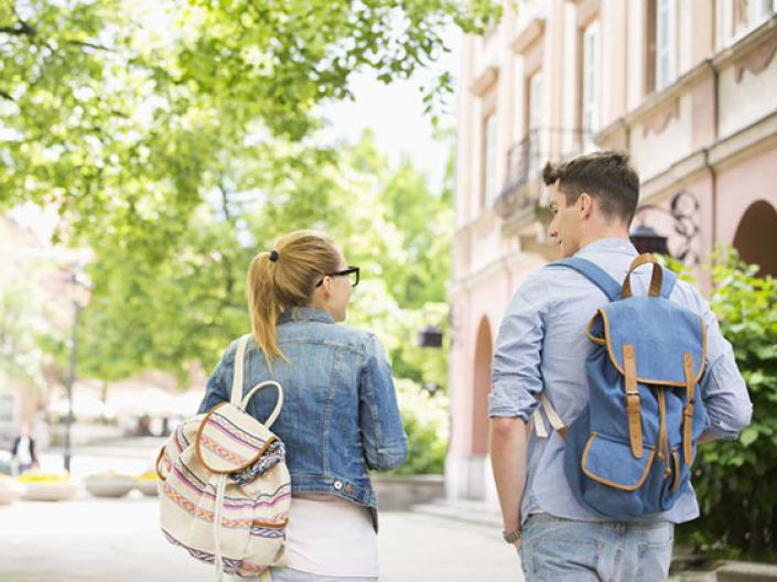 Image of two students walking away, with their backs to the camera, one wearing a jean jacket and white canvas backpack with brown and grey stripes and glasses, the other wearing a light blue shirt, jeans, and a denim fabric backpack with brown straps.  They are in the sun approaching a bright green tree on their left and a beige building on their right.  They are on a white path.