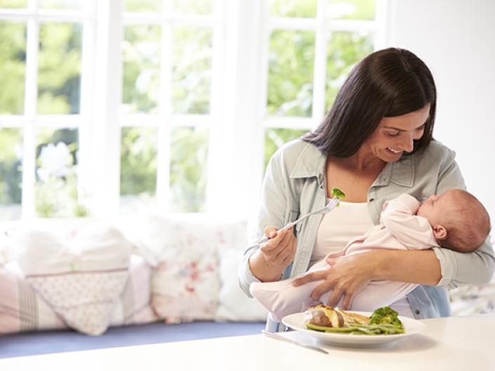 Image of mother holding baby with a plate of food in front of her on a large white counter, with brocolli a white meat on the plate. the mother is holding a fork with a piece of brocolli on it and smiling down at the baby, with a large window and couch with white and pink patterned cushions behind her