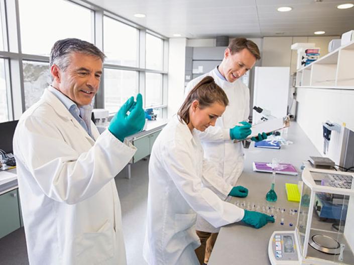 Image of three medical professionals in a lab setting, in white lab coats and green latex gloves, working with small samples spread out on a table on the right with a medium sized lab machine and other small lab equipment with bright windows to the left and some shelving above the counter to the right