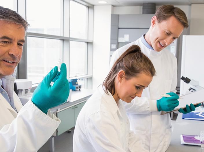 Image of three lab professionals in white lab coats and green latex gloves, the first holding up a test tube and looking at it, the second looking down at something outside the frame in front of her, the third holding a clipboard looking over the shoulder of the second and writing on the clipboard. they are in a lab environment with bright widows to the left and 3 medium sized pieces of equipment, one a microscope, with white shelves and miscellaneous items on the right.
