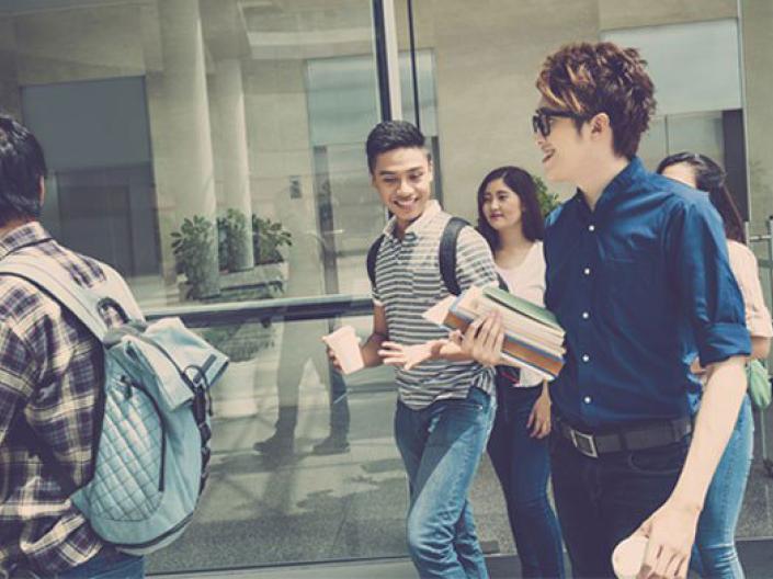 Image of five students walking in the sun in front of a large glass window. they are wearing jeans. two are wearing backpacks, one is holding a cup, and one, wearing sunglasses, is carrying books in one hand and a cup in the other. The two other students are walking in the background, obscured by the three in the foreground