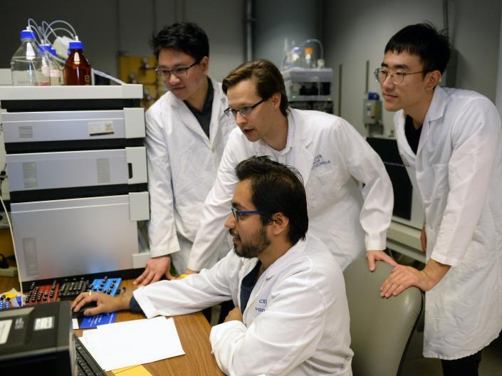 Four men in white lab coats gather around a computer. One is sitting and the other three are standing.