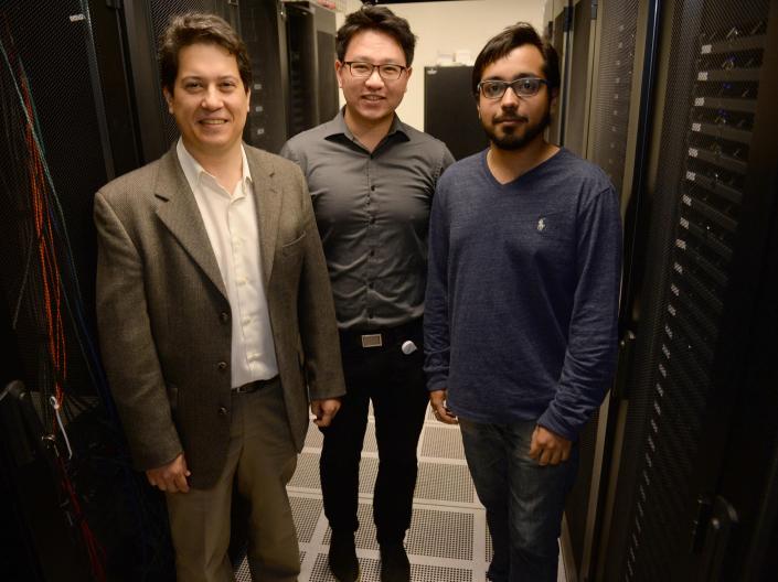 Three men stand next to each other in an aisle of a server room.