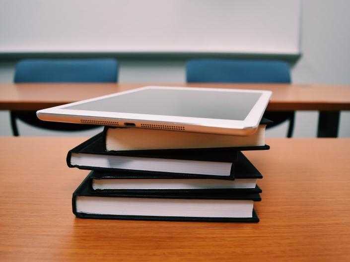 A stack of books with a tablet on top sitting on a wooden table in a classroom.