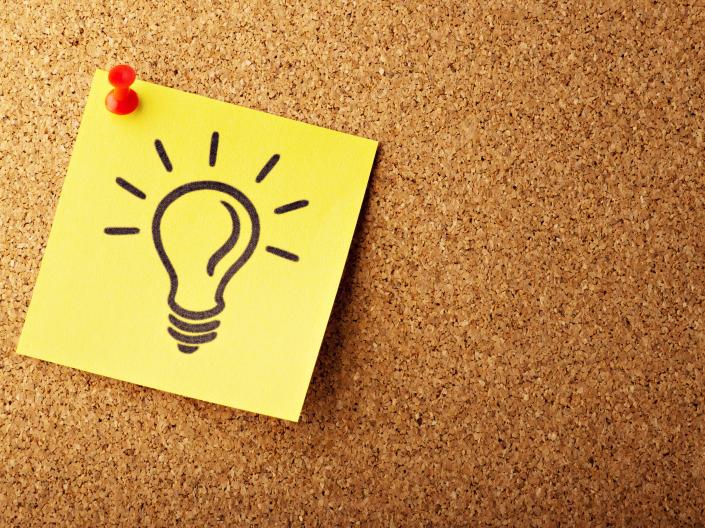 A yellow sticky note with a lightbulb drawn on it pinned to a cork board.