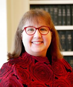 Portrait of Tammy Randall wearing a red shirt and red glasses sitting in front of a book case.