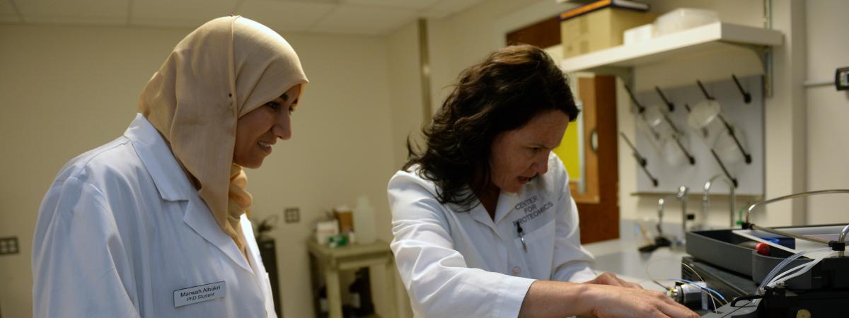 Two women wearing white coats work with scientific equipment.