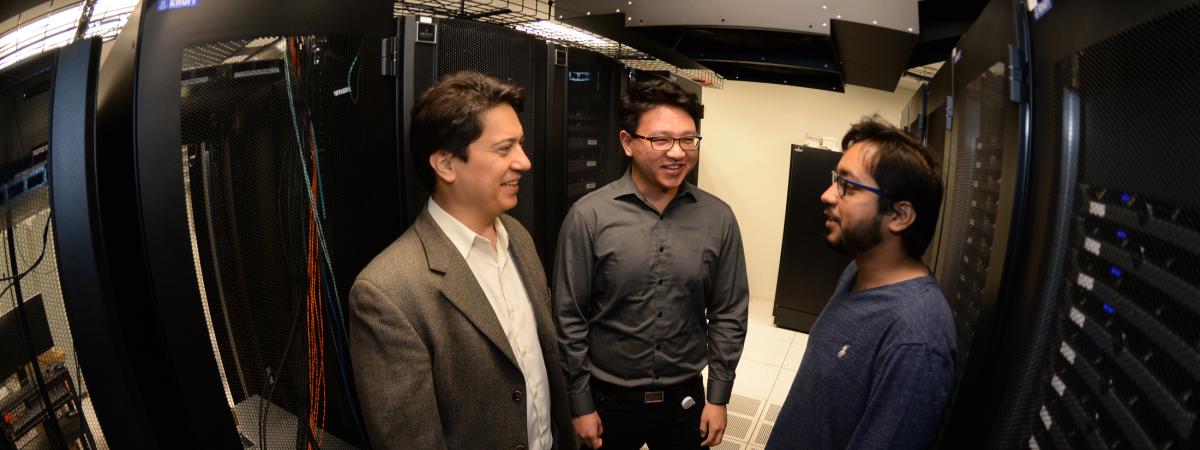 Three men stand in an aisle of a server room while talking and smiling.