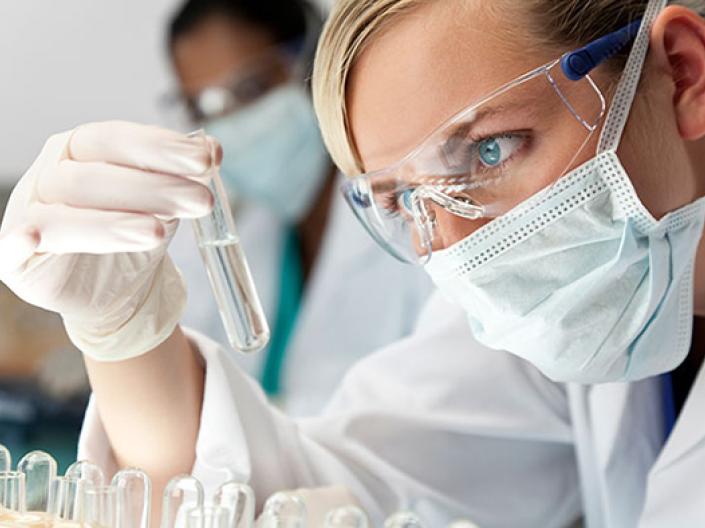 Image of lab professional with safty glasses, surgical mask, white lab coat and white latex gloves, analysing  a clear test tube with a clear liquid, with test tubes in a rack, some upside down, in front of her.  There is another lab professional dressed similarly in the background and a microscope in the midground to the left.