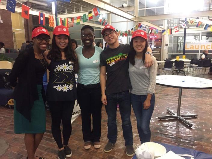 Image of five students, four of whom are wearing red hast with visors, from Case Western Reserve University's culture-based clubs in a student area decorated for Student Dietetic Association's Multicultural Food Day with small flags of many countries decorating the ceiling, with small round white tables, black chairs and many windows in the background, with spoons plates and napkins seen in the bottom right foreground
